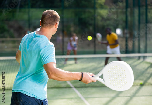 Rear view of man playing paddle tennis match on outdoor court on blurred background of opponents. Sport and active lifestyle concept