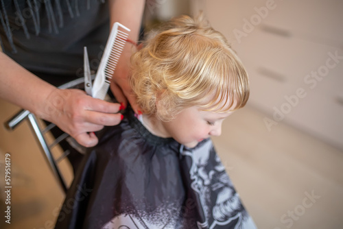 A hairdresser cuts a little girl at home in the middle of a room. Haircut of children at home
