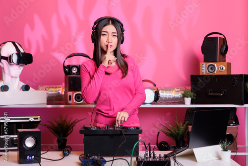 Asian artist putting forefinger on lips gesturing shh sign while performing electronic music in studio over pink background. Musical performer playing sounds to produce melody at mixer console photo