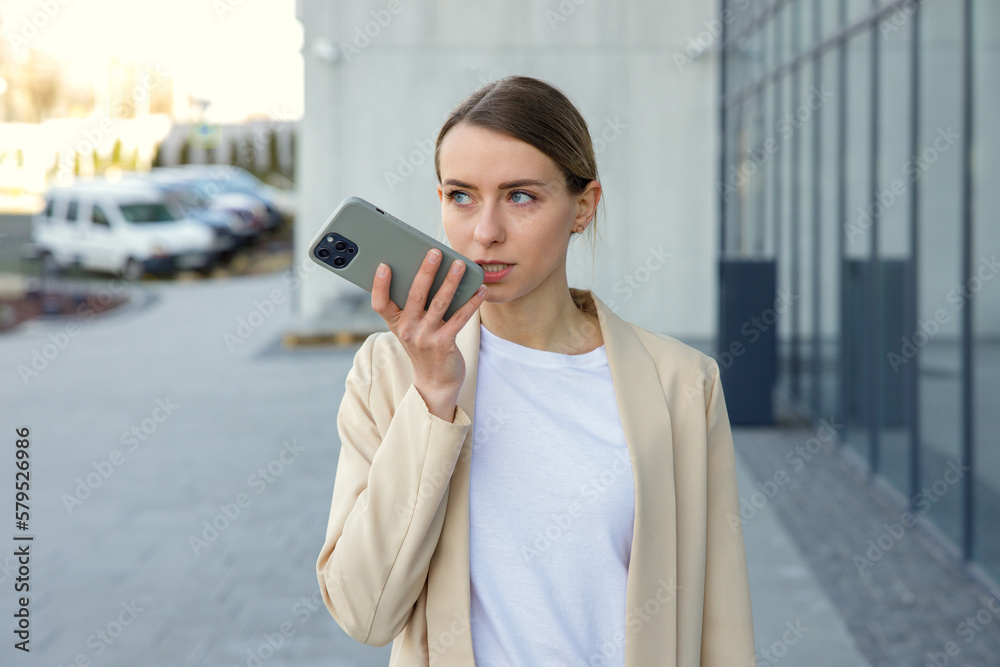 Smiling woman with smartphone in hand recording voice message to colleague outdoors. Smiling female employee in official wear walking near business center. Smiling woman with smartphone in hand recording voice message to colleague outdoors. Smiling female employee in official wear walking near business center.
