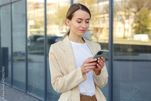 Smiling caucasian woman surfing internet on modern smartphone while walking outdoors. Charming female blonde using mobile near office building.
