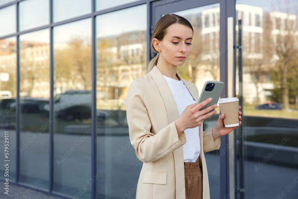 Busy caucasian business woman walking, using smartphone and holding coffee cup on street. Attractive female typing message near office building during coffee break.