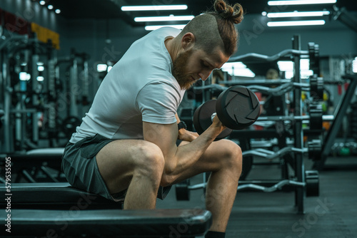 A man in sportswear sitting on the gym bench and workout on his arms with dumbbells. 