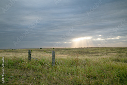 Rays of Sun Shine Through Cloudy Skies in a Flat Grass Field with Wooden Fence in Foreground