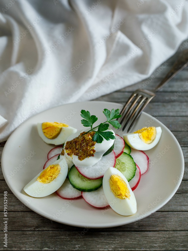 Plate with healthy spring vegetarian salad with radish, cucumber, egg and parsley with sour cream. and mustard, top view on wooden background with fork and napkin, russian summer food. Vertical