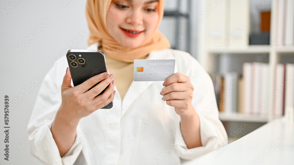 Cropped image of beautiful young Asian Muslim woman holding her phone and credit card. Cropped image of beautiful young Asian Muslim woman holding her phone and credit card.