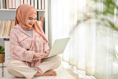 Beautiful young Asian Muslim woman wearing hijab using her laptop in her living room