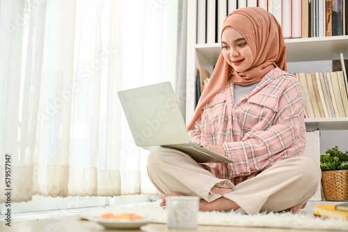 Attractive Asian Muslim female working from home, using laptop in her living room.