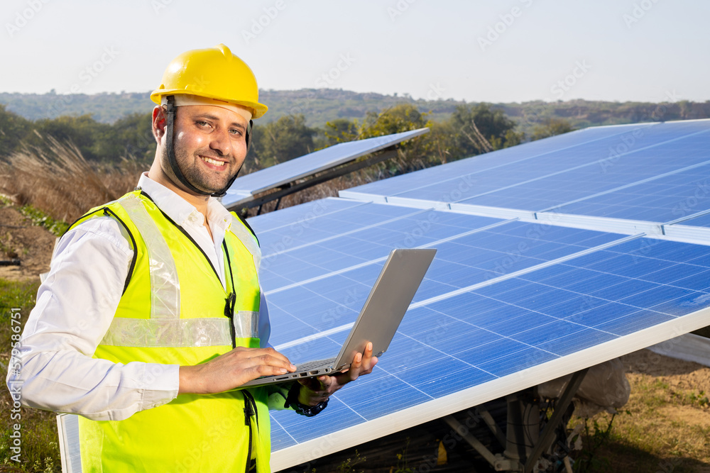 Portrait of Young indian man technician wearing yellow hard hat holding laptop standing near solar panels.Industrial worker solar system installation, renewable green energy generation concept. Portrait of Young indian man technician wearing yellow hard hat holding laptop standing near solar panels.Industrial worker solar system installation, renewable green energy generation concept.