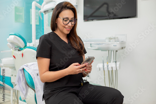 Young Latina dentist sitting in her office, checking her cell phone