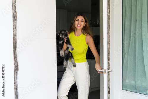 Welcome. Portrait of cheerful woman standing in doorway of modern apartment, greeting visitor and inviting guest to enter her home, holding a puppy