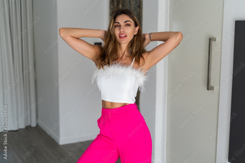 Stylish fit tanned beautiful woman in fashion pink pants and top posing in bedroom at home, preparing for party, dressing up Stylish fit tanned beautiful woman in fashion pink pants and top posing in bedroom at home, preparing for party, dressing up