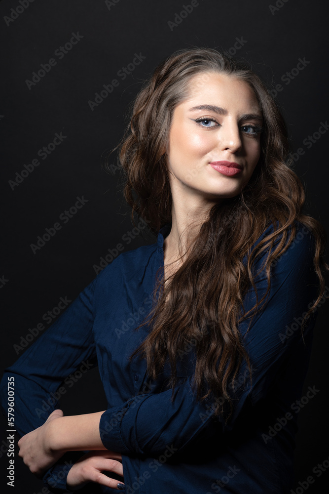Makeup and fashion concept. Brunette woman with long wavy hair sensual studio portrait. Model wearing blue blouse and looking at camera with blue eyes and seductive look. Dark studio background