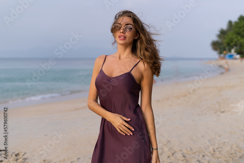 Stylish romantic tender sensual woman in silk dress and sunglasses on the beach at sunset