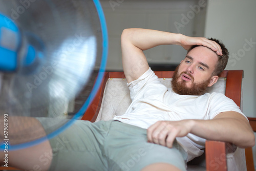 Young bearded man using electric fan at home, sitting on couch cooling off during hot weather, suffering from heat, high temperature photo