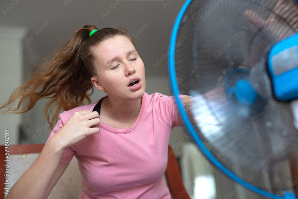 Young woman using electric fan at home in living room, sitting on couch