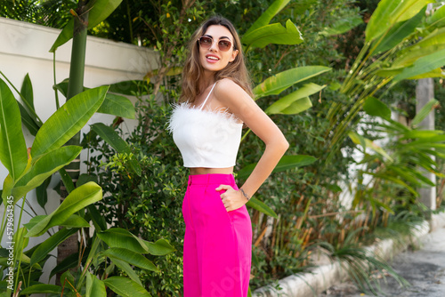 Stylish fit tanned beautiful woman in sunglasses  fashion pink pants and white top posing outdoor at luxury tropical villa