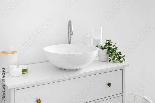 Sink with bath accessories, candles and houseplant on chest of drawers near light wall