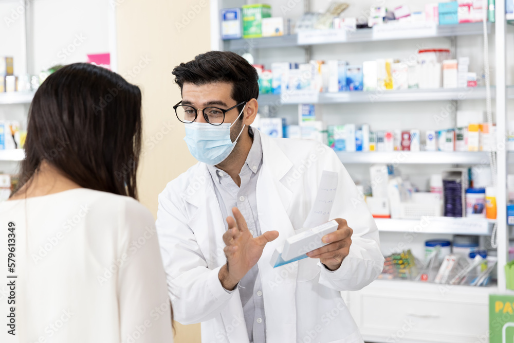 Middle eastern male pharmacist wearing protective hygienic mask to prevent infection selling medications to woman patient to prescription and making drug recommendations in modern pharmacy Middle eastern male pharmacist wearing protective hygienic mask to prevent infection selling medications to woman patient to prescription and making drug recommendations in modern pharmacy