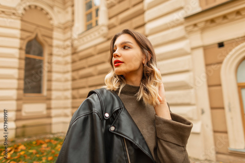 Stylish beautiful fashionable woman model with red lips in fashion trendy clothes with a leather jacket walks in the autumn town near vintage building