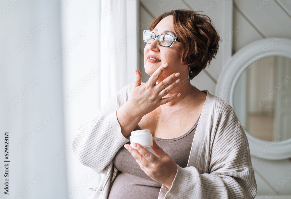 Smiling middle aged plus size woman with day cream near window at home