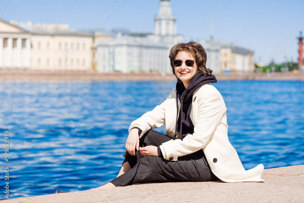 Girl sits on concrete flooring embankment in white coat and black sweatshirt in sunglasses, smiles happily on a warm sunny day. Relax in the city