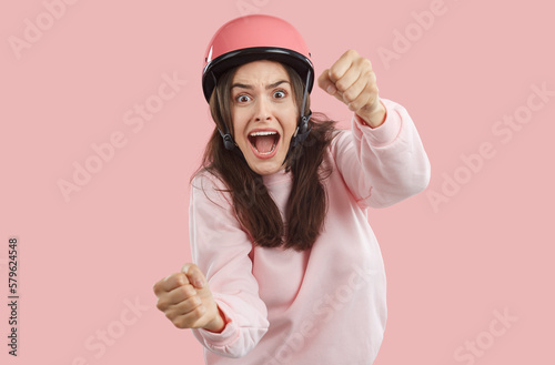Scared afraid young brunette woman with opened mouth in pink helmet and sweatshirt imagines she is driving car and holding steering wheel on pink background. Human emotions, driving a car concept.