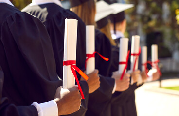 College or university graduates at outdoor graduation ceremony. Diverse group of students standing in row and holding diploma scrolls tied with red ribbons. Concept background. Cropped closeup shot