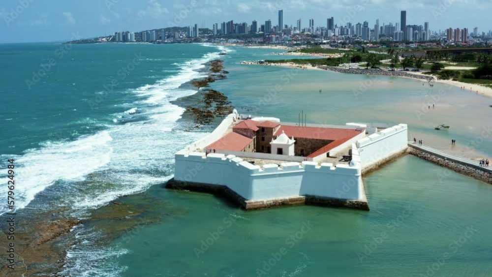 Dolly in aerial drone shot of the historic star shaped Reis Magos fort built on a reef with the coastal capital city of Natal in Rio Grande do Norte, Brazil in the background on a warm summer day