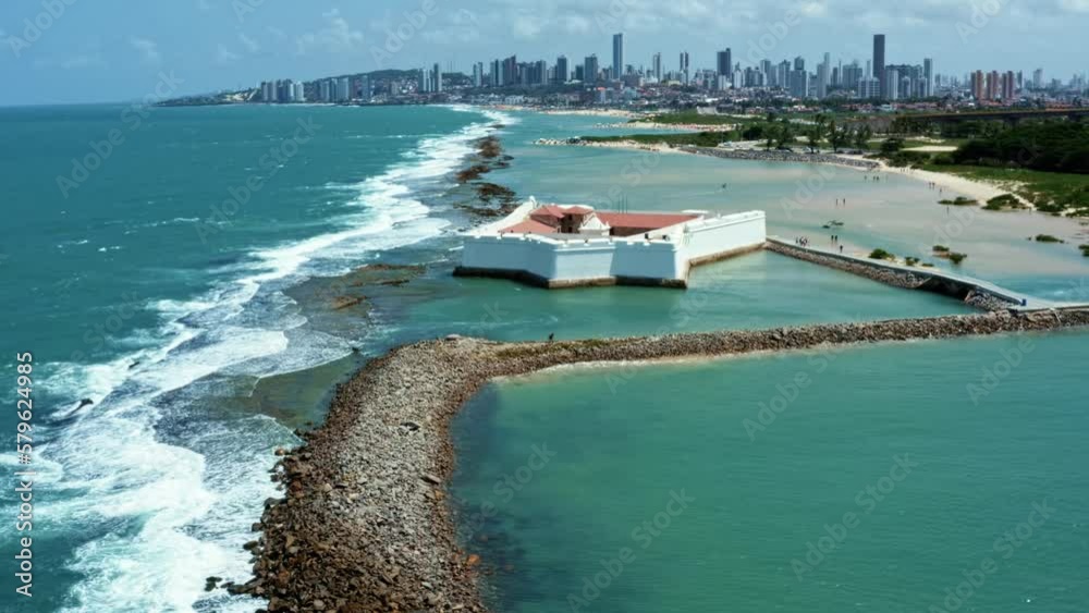 Dolly in aerial drone wide shot of the historic star shaped Reis Magos fort built on a reef with the coastal capital city of Natal in Rio Grande do Norte, Brazil in the background on a warm summer day