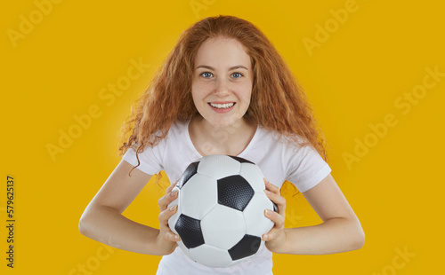 Soccer fan happy smiling readhead woman with curly long hair wearing white t-shirt holding football ball in hands on yellow background and looking at camera. Victory, goal, soccer game, fan concept. © Studio Romantic