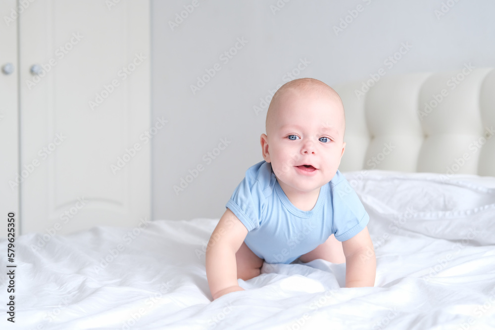 smiling boy with big blue eyes in a bodysuit learns to crawl on white bedding. Healthy newborn baby