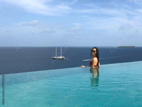A beautiful brunette woman with long hair stands with her back to the roof of a house in a pool overlooking the ocean in the Maldives. View from a drone.