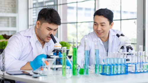 Asian professional male scientist researcher in white lab coat and rubber gloves sitting using microscope inspecting quality of vegetable in laboratory with colleague