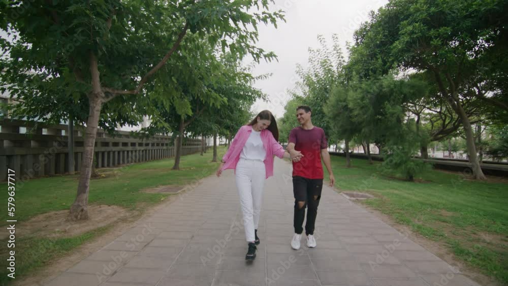 Young teen caucasian couple walking holding hands in city green park. Date in summer park. Attractive girl happily whirling fit boy. Boyfriend and girlfriend enjoying spend time in love together. 4k Young teen caucasian couple walking holding hands in city green park. Date in summer park. Attractive girl happily whirling fit boy. Boyfriend and girlfriend enjoying spend time in love together. 4k