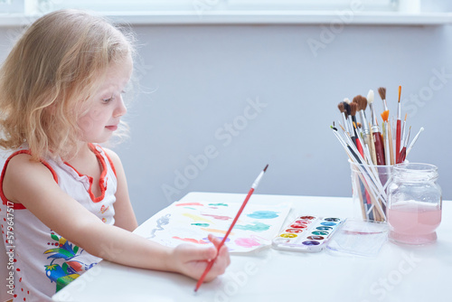 A curly-haired cute girl paints at a small table in kindergarten. photo