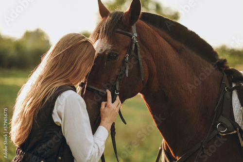 Young beautiful female jockey strokes and hugs the horse's head and prepares for the competition. Jumping training in the meadow in summer evening.
