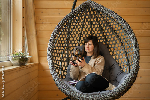 Remote work and escaping to nature concept. Woman sit in egg chair swing and holds a vase of cones in wooden tiny cabin house. photo