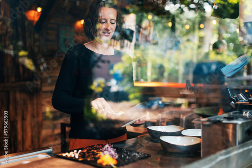 Female chef at a barbecue grill house restaurant preparing dishes with colorful vegetable garnishes on black stone plates - Reflection on the kitchen glass