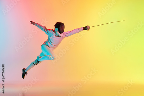 Long attack. Young man, male fencer with sword practicing in fencing over gradient pink-yellow background in neon light. Sportsman shows fencing technique photo