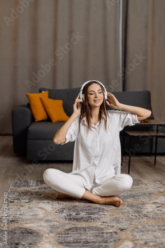 Happy young caucasian woman sitting on the floor at home, listening music by smartphone with headphones and dancing 