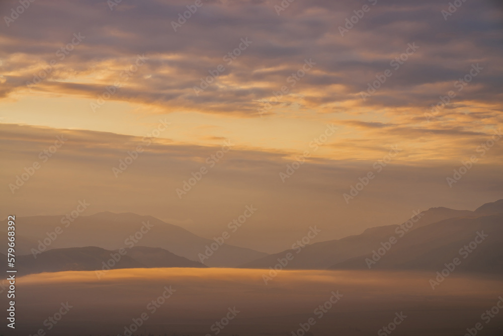 Scenic dawn mountain landscape with golden low clouds in valley among mountains silhouettes under cloudy sky. Vivid sunset or sunrise scenery with low clouds in mountain valley in illuminating color.