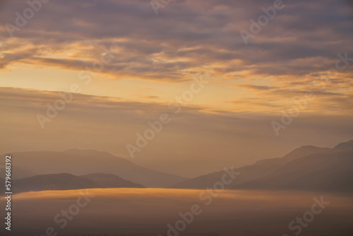 Scenic dawn mountain landscape with golden low clouds in valley among mountains silhouettes under cloudy sky. Vivid sunset or sunrise scenery with low clouds in mountain valley in illuminating color.