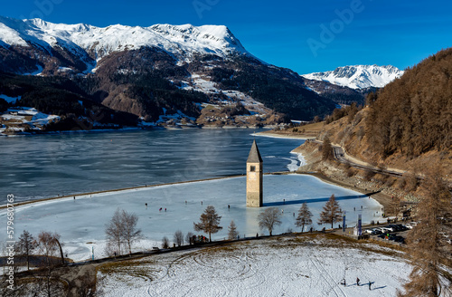 CURON VENOSTA, 06 January 2023: Tourists walk on the frozen lake of Resia. It is an artificial reservoir in the west of South Tyrol at Curon in Val Venosta, Italy
