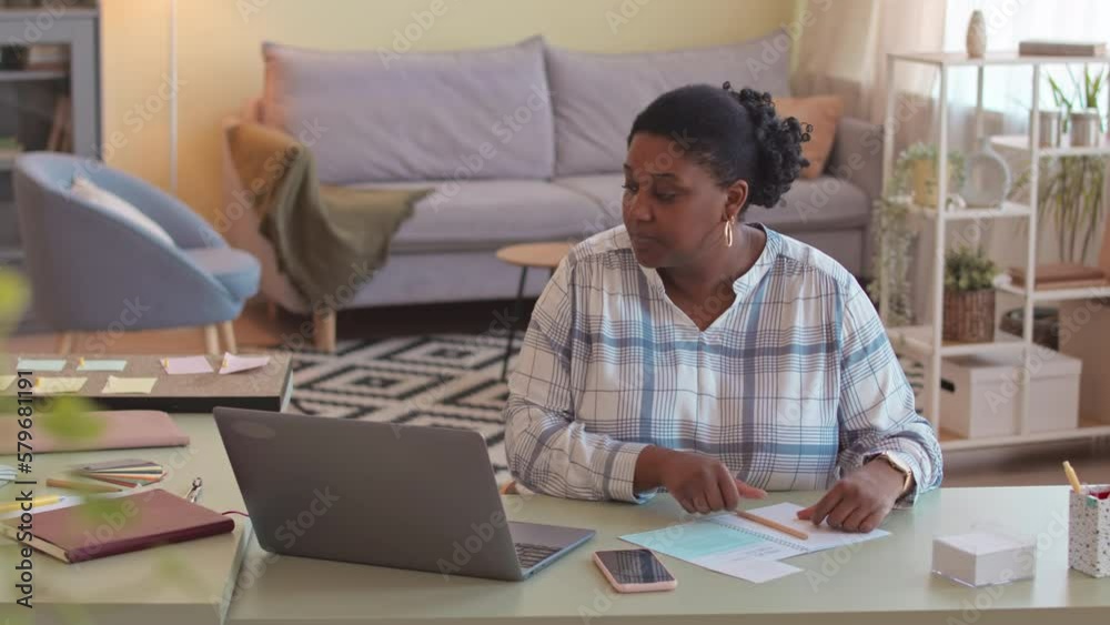 Waist up of young plus size African American woman working freelance at home, sitting in front of laptop in modern living room and looking through notes in notebook Waist up of young plus size African American woman working freelance at home, sitting in front of laptop in modern living room and looking through notes in notebook
