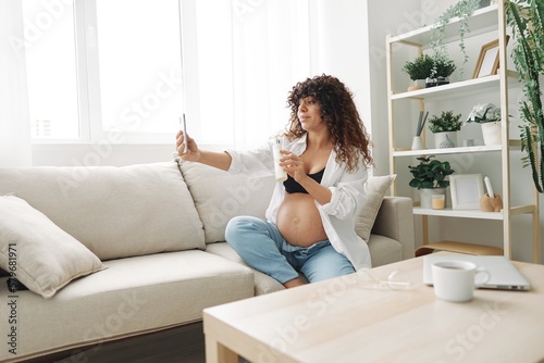 A pregnant woman blogger advertises a cream for stretch marks on the body during pregnancy, filming herself on the phone while sitting on the couch at home