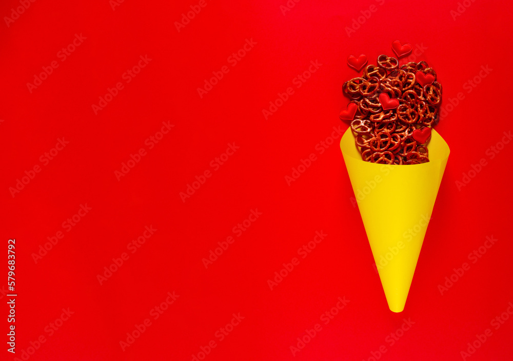 Mini pretzels and small colorful hearts in yellow paper cone lie on red background. Pretzel sunday concept. Banner with blank space for text Mini pretzels and small colorful hearts in yellow paper cone lie on red background. Pretzel sunday concept. Banner with blank space for text
