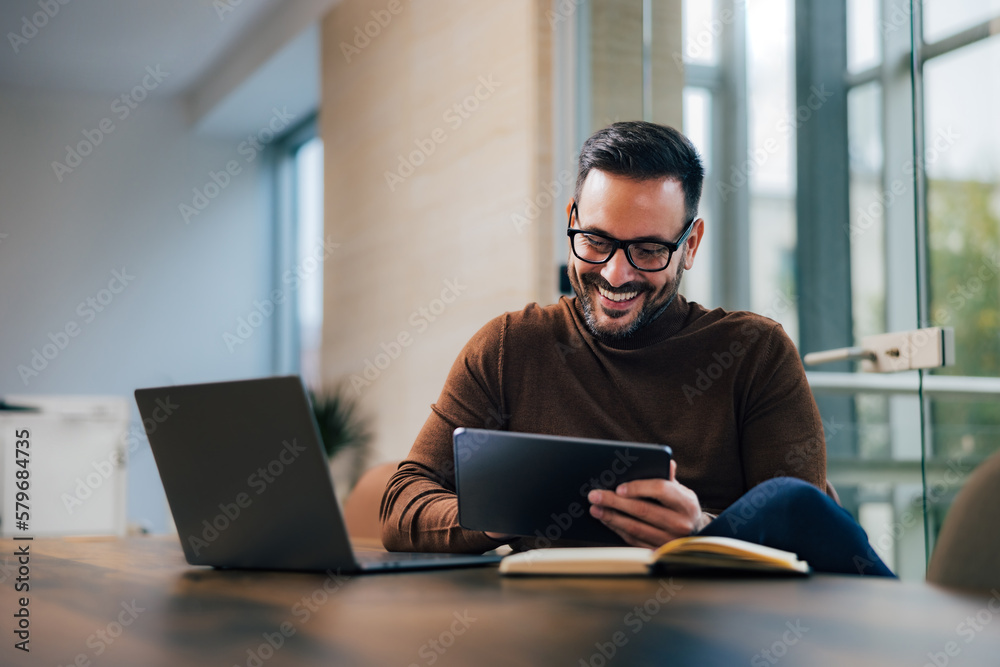 Smiling businessman using a tablet, sitting in front of the laptop at the office, casually dressed. Smiling businessman using a tablet, sitting in front of the laptop at the office, casually dressed.