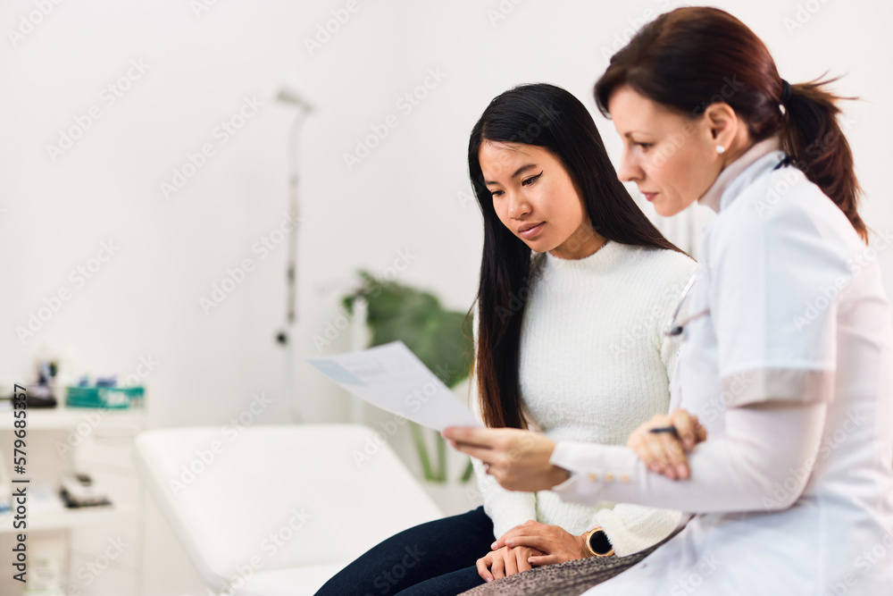 A confident female doctor sitting with an Asian patient, holding a result. A confident female doctor sitting with an Asian patient, holding a result.