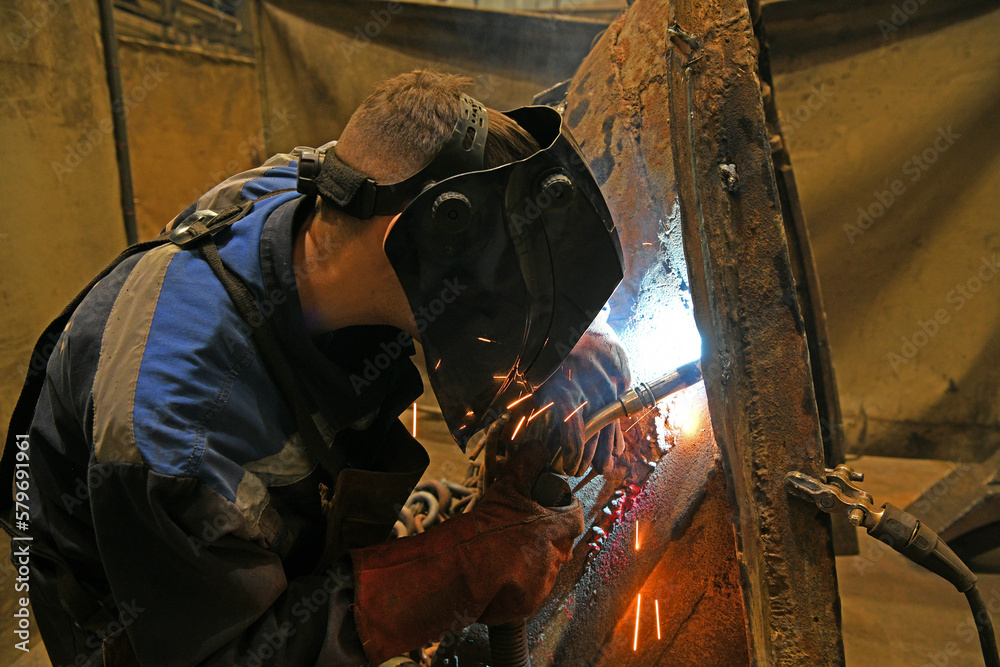 A worker welding metal parts on a construction site. A welder welds parts of a large machine in a metallurgical workshop. An interesting example of manual work.	